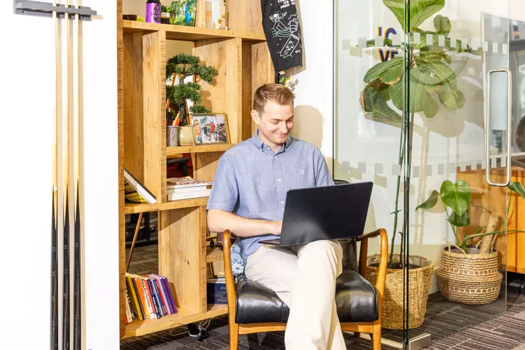 A man sits in an office chair, smiling whilst working on a laptop. He is in a bright, modern space with wooden shelves, books, plants, and glass walls.