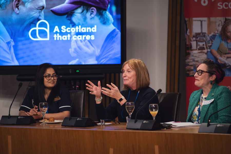 Three women sit at a panel table with microphones and drinks, engaged in discussion. A screen behind them displays the text “A Scotland that cares” alongside an image of two people smiling.
