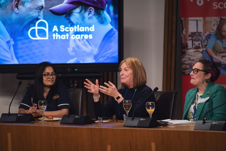 Three women sit at a panel table with microphones and drinks, engaged in discussion. A screen behind them displays the text “A Scotland that cares” alongside an image of two people smiling.