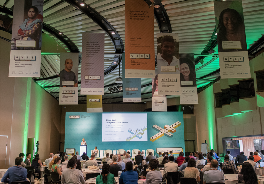 A conference hall with attendees seated at round tables, a person speaking on stage, and large banners featuring portraits and biographies hanging from the ceiling, under green and white lighting.