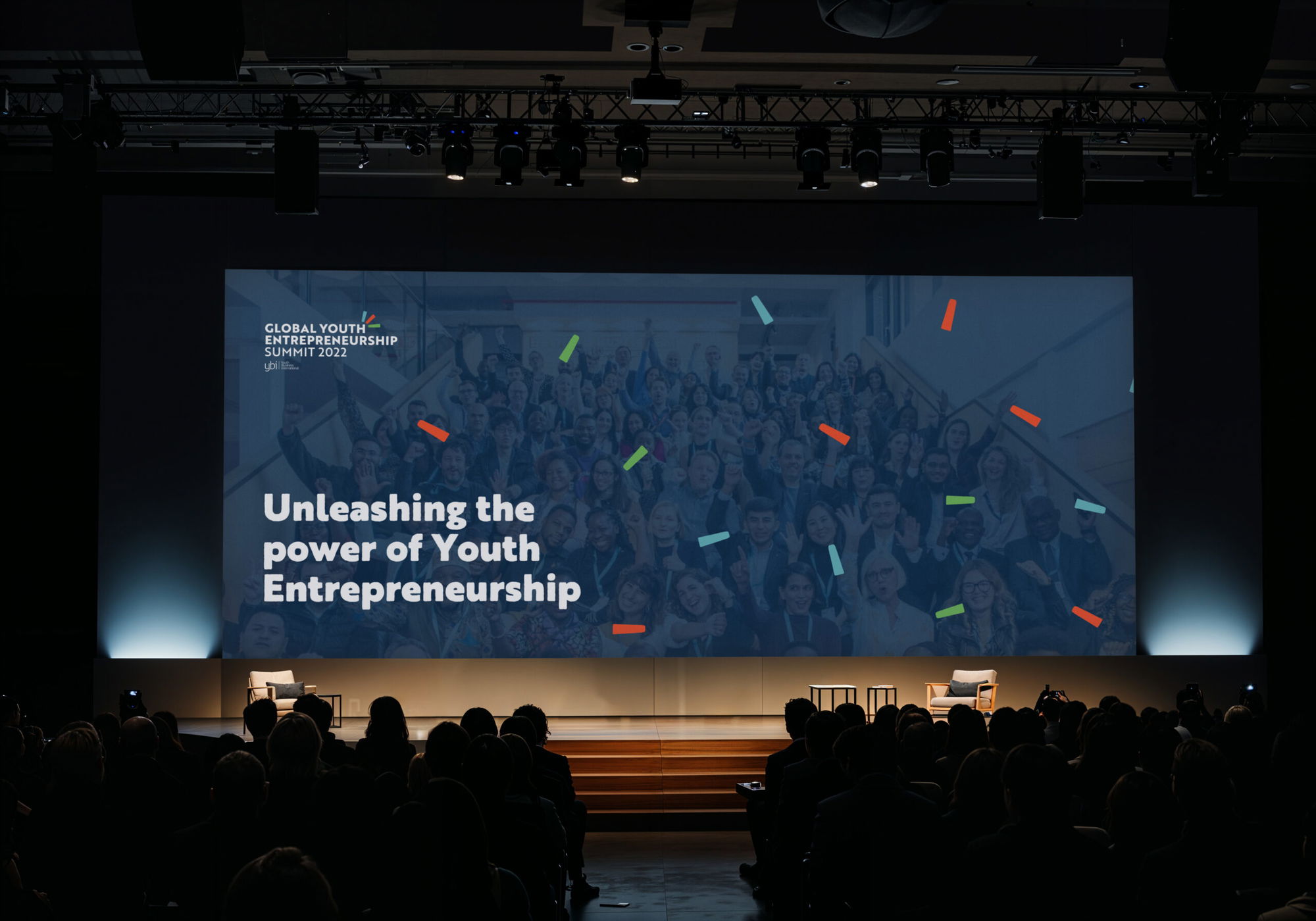 A large screen at an event displays Global Youth Entrepreneurship Summit 2022 with sponsor logos. A woman speaks into a microphone in the background. The audience is seated and watching.