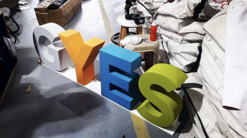 Large 3D letters spelling YES in grey, orange, blue, and green stand on a workshop floor surrounded by tools, stools, cardboard, and white bags.