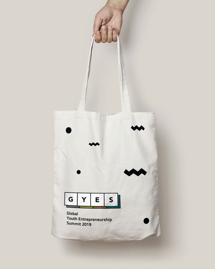 A hand holds a white tote bag with abstract black shapes and the text “GYES Global Youth Entrepreneurship Summit 2019” printed on it in black and coloured fonts.