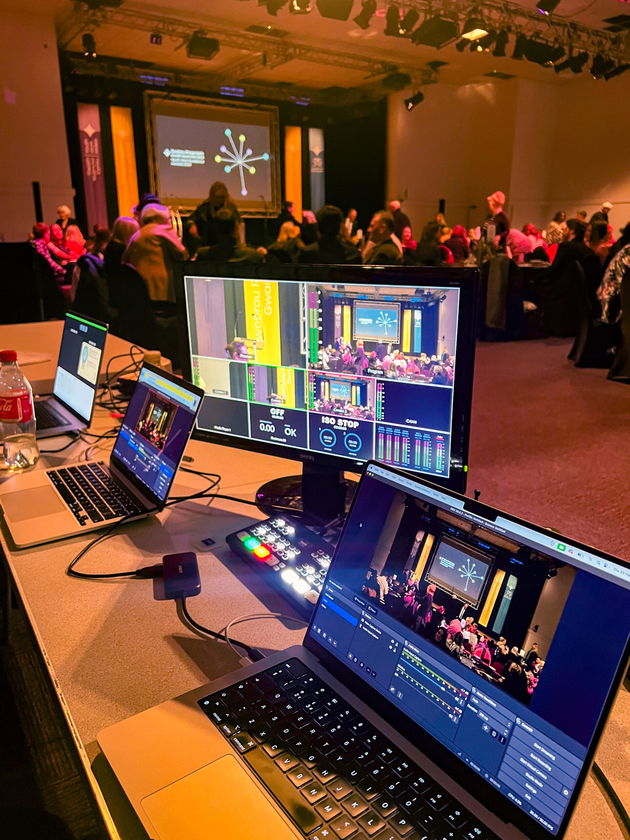 A control desk with monitors and laptops displaying a live event feed is set up in the foreground at a conference, with an audience seated and a presentation happening on stage in the background.