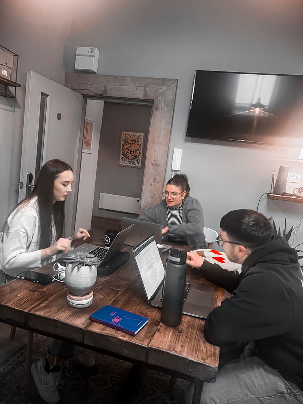 Three people are sitting round a table with laptops and papers, appearing to work or collaborate in a modern office space. A TV is mounted on the wall in the background.