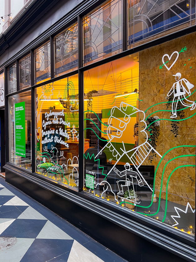 Shopfront window with colourful, hand-drawn illustrations and text, featuring figures, hearts, and abstract lines. Warm yellow light glows from inside, and chequered flooring is visible outside.