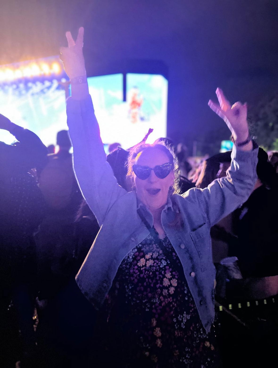 A woman in sunglasses and a denim jacket raises her hands in excitement at an outdoor concert at night, with bright stage lights and a large screen in the background.