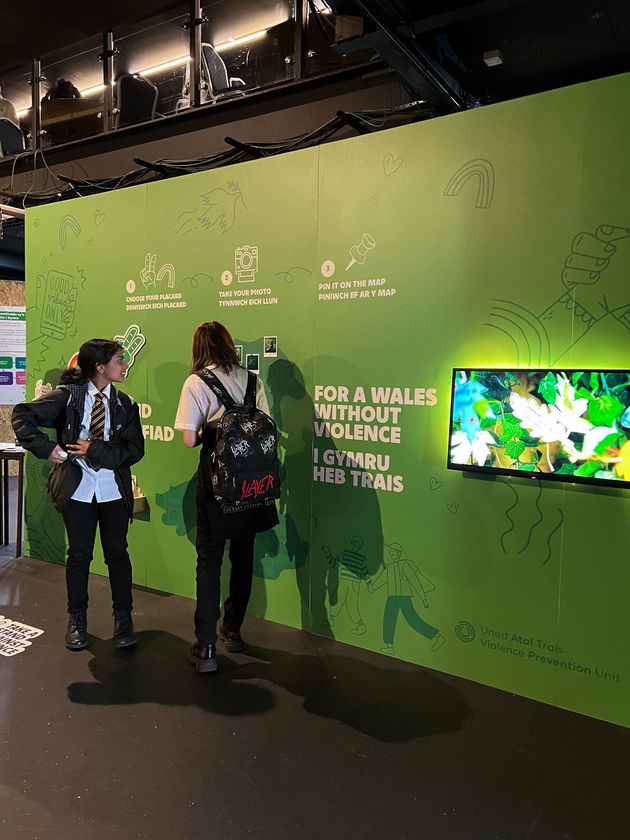 Two students in school uniforms stand in front of a green wall display about preventing violence in Wales, featuring bilingual text, instructions, and a mounted screen with nature imagery.