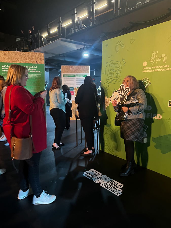 People stand in a queue at an indoor event with green walls featuring text and graphics. A woman holds a large hand-shaped sign, while others look at their mobiles or interact with displays. The setting is modern and dimly lit.