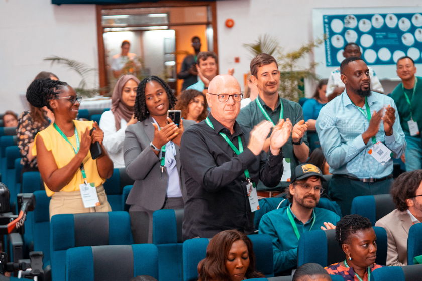 A diverse group of people stands and applauds in an auditorium, many wearing green lanyards and name badges, suggesting a conference or seminar setting. Some are smiling, whilst others are taking photos.