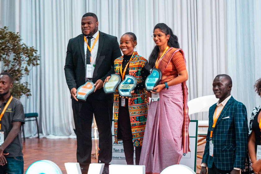 Four young adults standing on a stage, three of whom are holding award plaques and smiling. They are dressed in formal and traditional attire, with lanyards round their necks. A white curtain and some greenery are in the background.