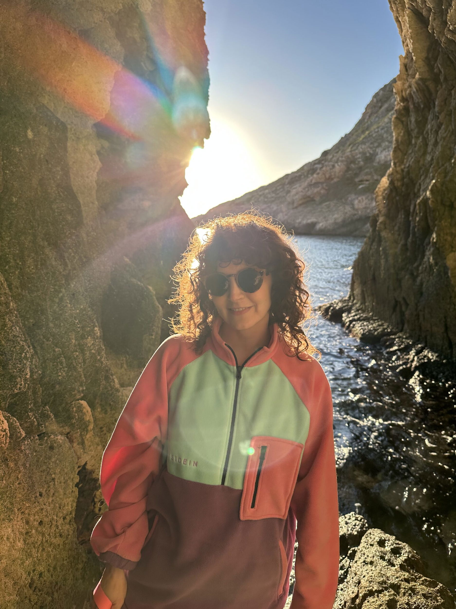 A person with curly hair and sunglasses stands between rocky cliffs near the water at sunset, wearing a colourful jacket with pink, green, and purple sections. Sunlight and lens flare create a vibrant effect in the image.