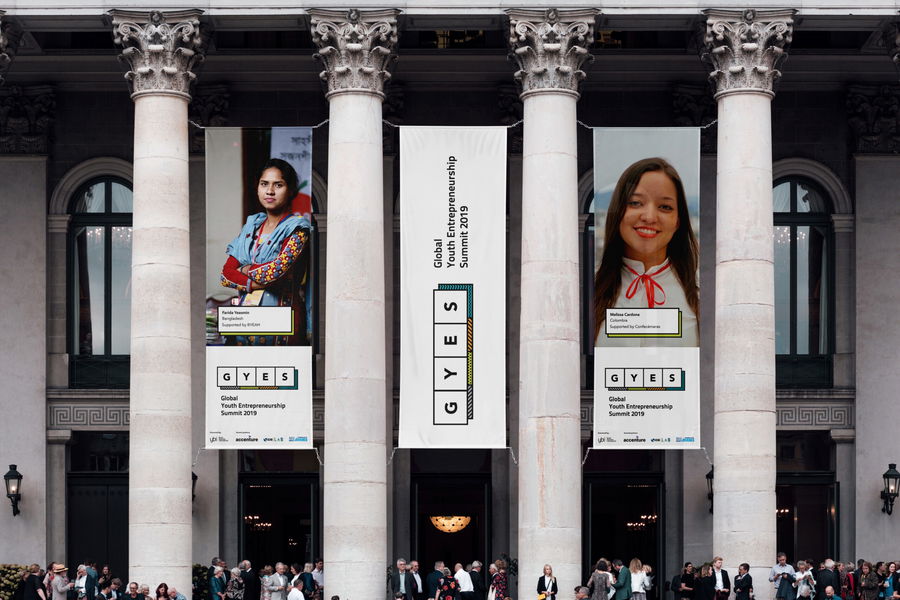 Large banners for the Global Youth Entrepreneurship Summit 2019 hang between tall columns on a grand building, displaying photos of two young women. People gather below the banners at the event entrance.