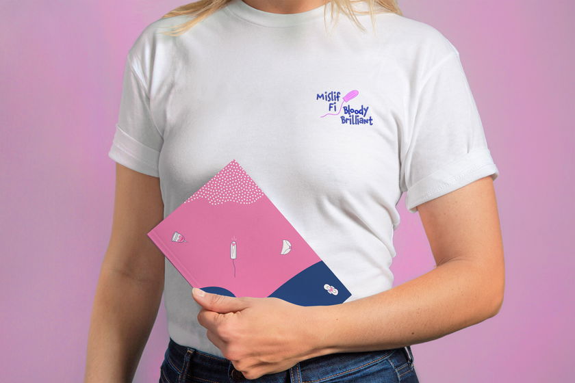 A person wearing a white T-shirt with the text Mislif Fi Bloody Brilliant holds a pink and blue notebook featuring illustrations of a tampon, pad, menstrual cup, and small dots.