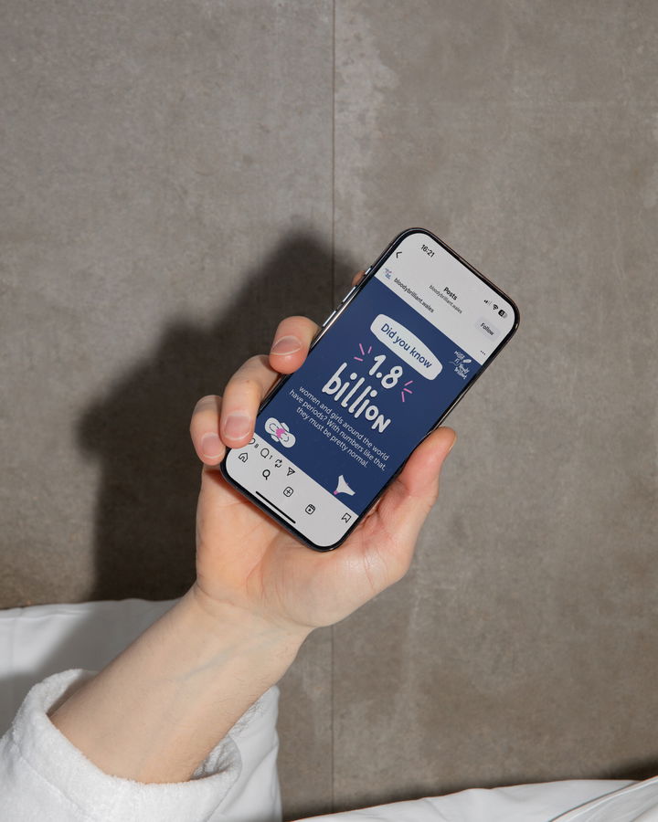 A hand holding a mobile phone, displaying an Instagram post that reads Did you know? 1.8 billion on a blue background, with social media icons visible below. The background is a plain, neutral wall.