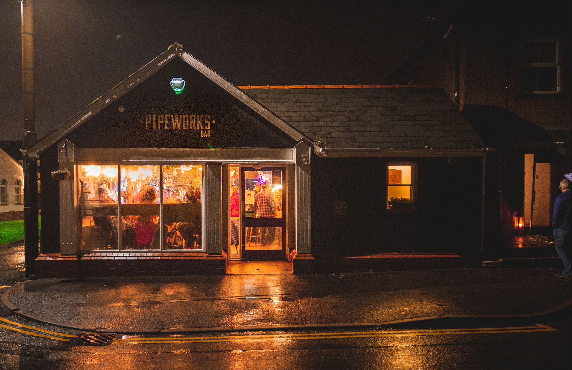 A cosy bar called The Pipeworks Bar glows warmly at night, with people visible inside through large windows. The street outside is wet, reflecting the light from the bar.