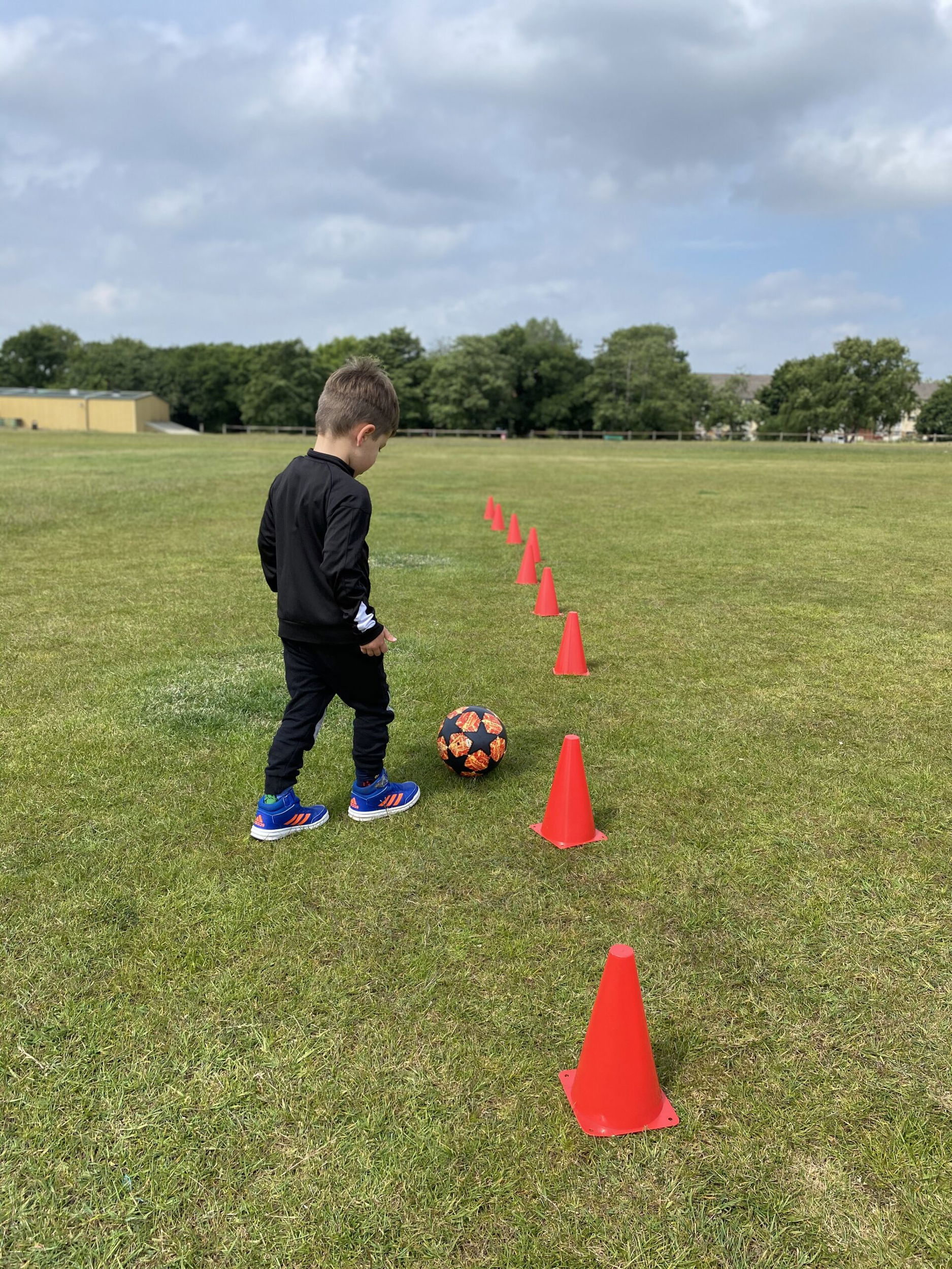 A young child in sports kit stands on grass, about to dribble a football through a line of red training cones on a pitch under a partly cloudy sky.