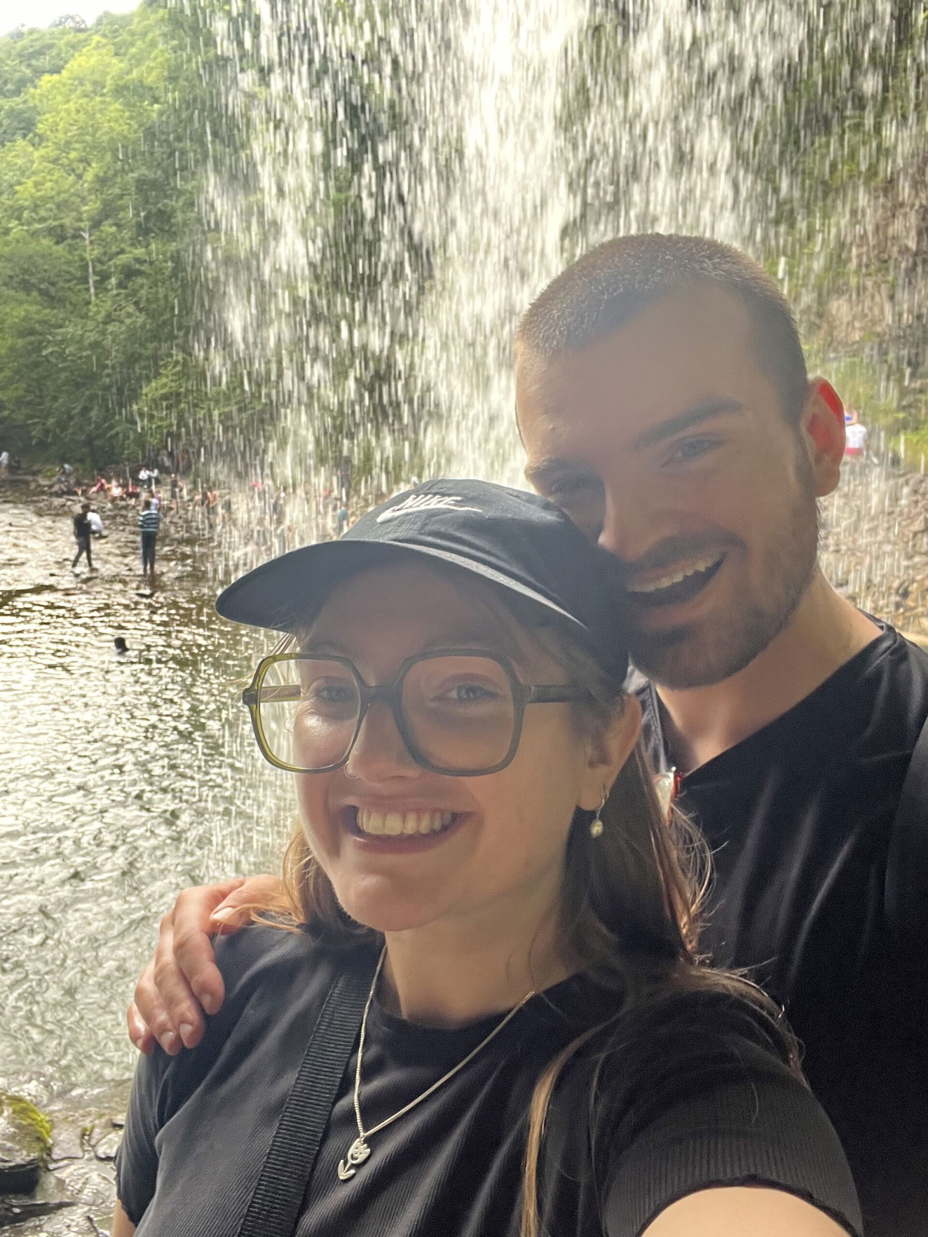 A smiling couple pose for a selfie in front of a waterfall, standing close together. The background shows flowing water, a pool, and several people enjoying the natural scenery. The woman wears glasses and a cap.