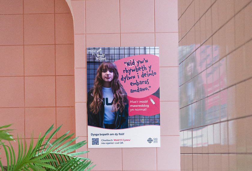 A poster on a tiled wall features a young woman with long hair and a serious expression. Welsh text appears in a speech bubble above her, and there are various logos and a QR code at the bottom of the poster.