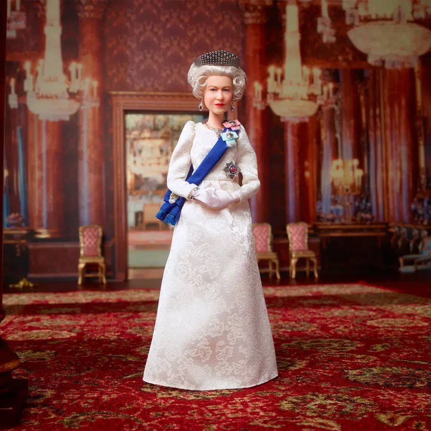 A doll dressed as a queen in a white gown, blue sash, and tiara stands in an ornate room with red carpets, chandeliers, and elegant chairs—a tribute to The Queen’s Platinum Jubilee.