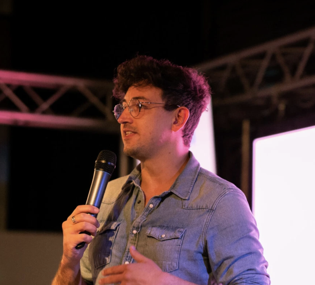 A person with curly hair and glasses, wearing a denim shirt, speaks into a microphone on stage under bright lights and metal truss structures, setting the perfect scene for an