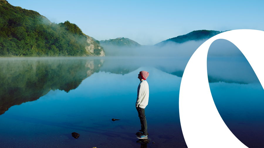 A person wearing a striped beanie stands by a calm lake, looking at misty hills reflected in the water, with a large abstract white shape on the right side of the image.