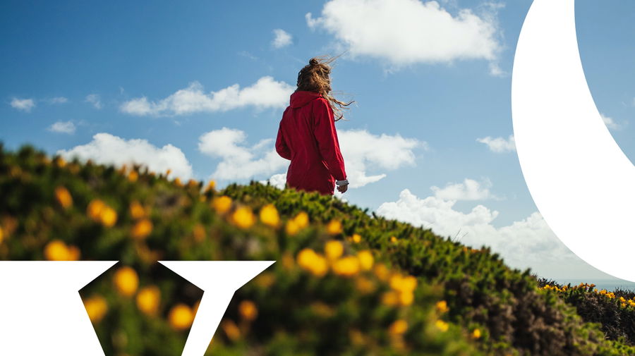 A person in a red jacket stands on a grassy hill with yellow flowers, looking at a blue sky with clouds. Large white graphic shapes overlay the image in the foreground.
