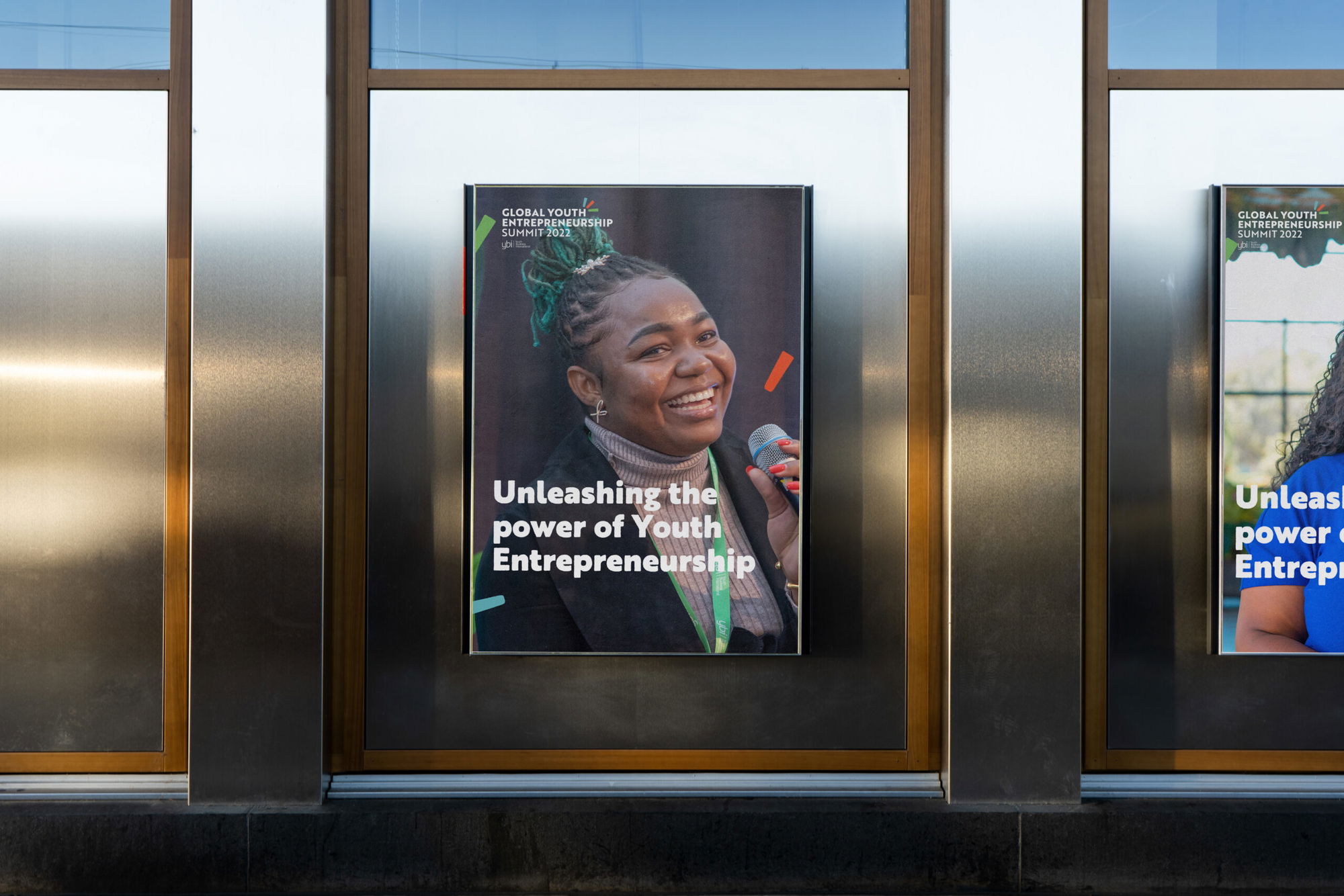 A poster on a wall shows a smiling woman holding a microphone with the text “Unleashing the power of Youth Entrepreneurship” and several sponsor logos at the bottom.