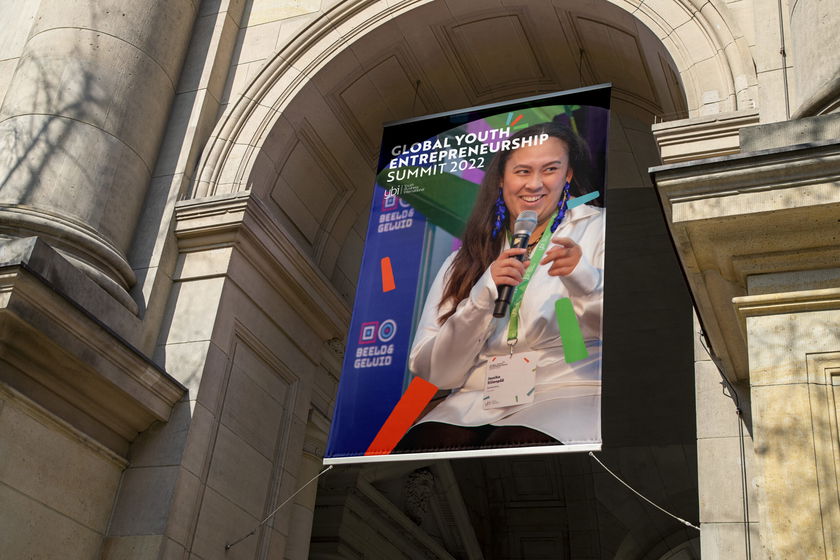 A banner for the Global Youth Entrepreneurship Summit 2022 hangs under a stone archway, featuring a smiling young woman holding a microphone. Logos of various sponsors are displayed at the bottom of the banner.