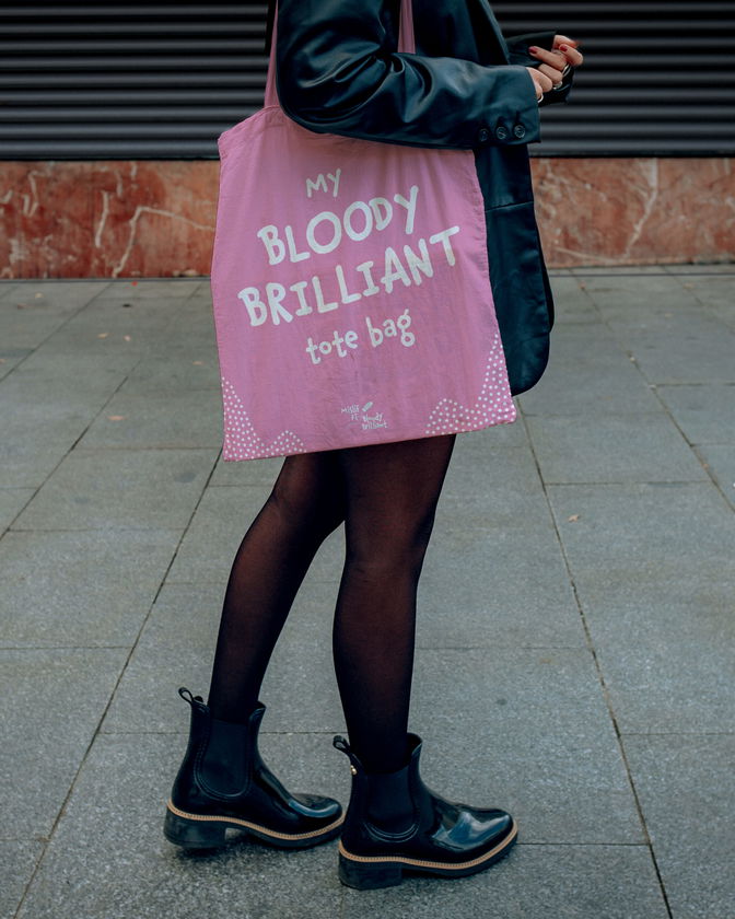 A person in a black jacket, black tights, and black boots carries a tote bag reading Bloody Brilliant! with pink and white designs, standing on a pavement.