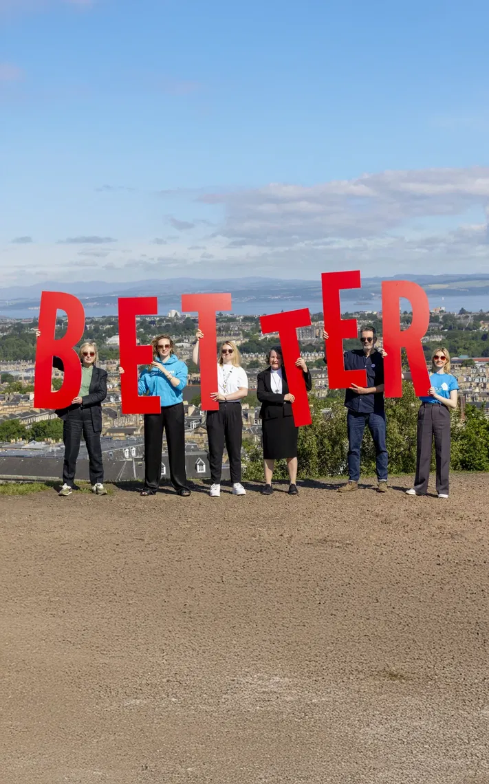 Six people standing outdoors on a hill, holding large red letters that spell BETTER against a backdrop of a city, trees, and blue sky with clouds.