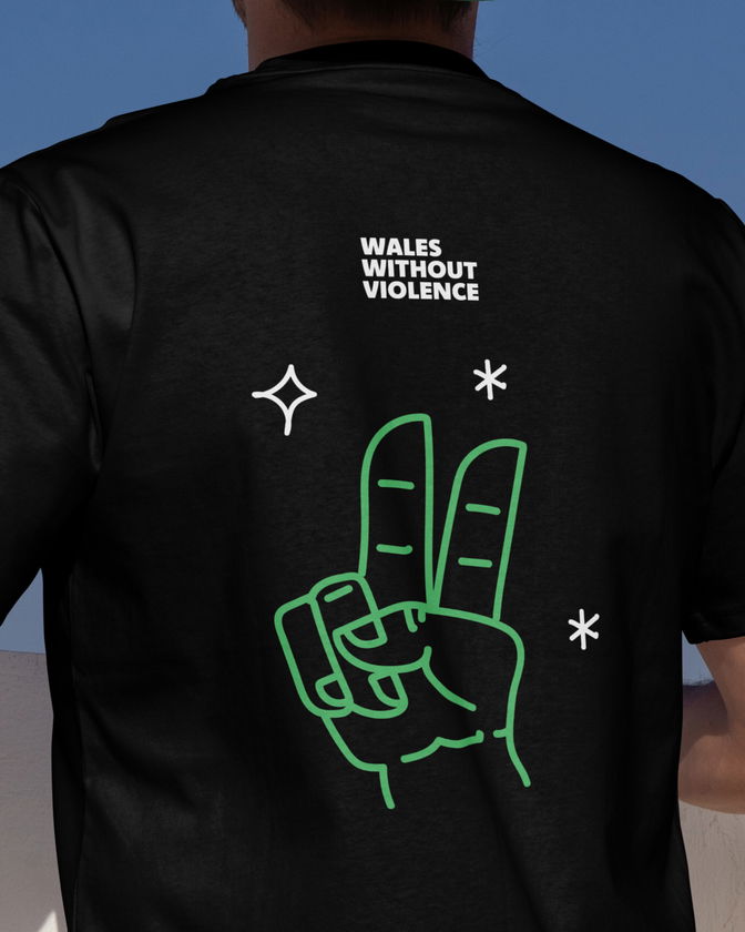 A person wearing a black shirt with WALES WITHOUT VIOLENCE printed on the back above a green outline of a hand making a peace sign, surrounded by white star illustrations.