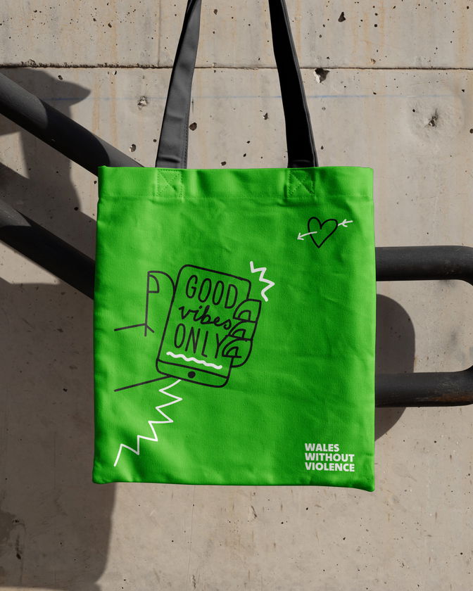 A bright green tote bag hangs on a black railing. It features a graphic of a mobile phone with the text Good vibes ONLY and doodle-style lines and a heart. The bottom corner reads WALES WITHOUT VIOLENCE in white text.