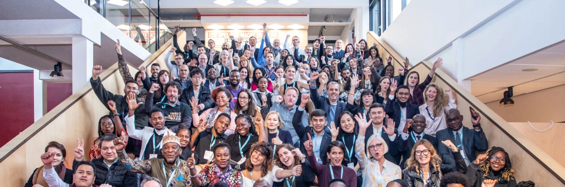 A large, diverse group of people sit and stand on wide indoor stairs, smiling and raising their fists in celebration or solidarity. The setting appears to be bright and modern, with glass balustrades and tall windows.
