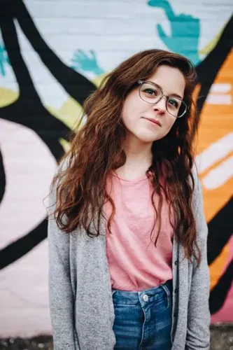 A young woman with long brown hair, wearing glasses, a pink shirt, grey cardigan, and blue jeans, stands in front of a colourful mural with abstract shapes and hands. She looks at the camera with a slight smile.