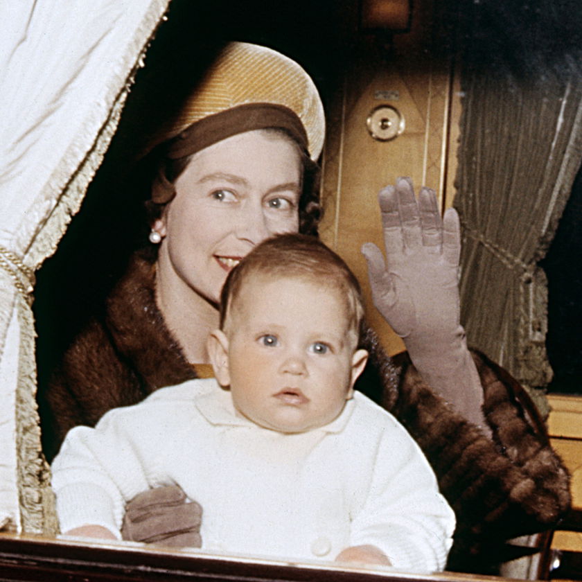 A woman wearing a hat, gloves, and fur coat smiles and waves from behind a curtain whilst holding a baby dressed in white on her lap, capturing the warmth and elegance of moments celebrated during The Queen's Platinum Jubilee.