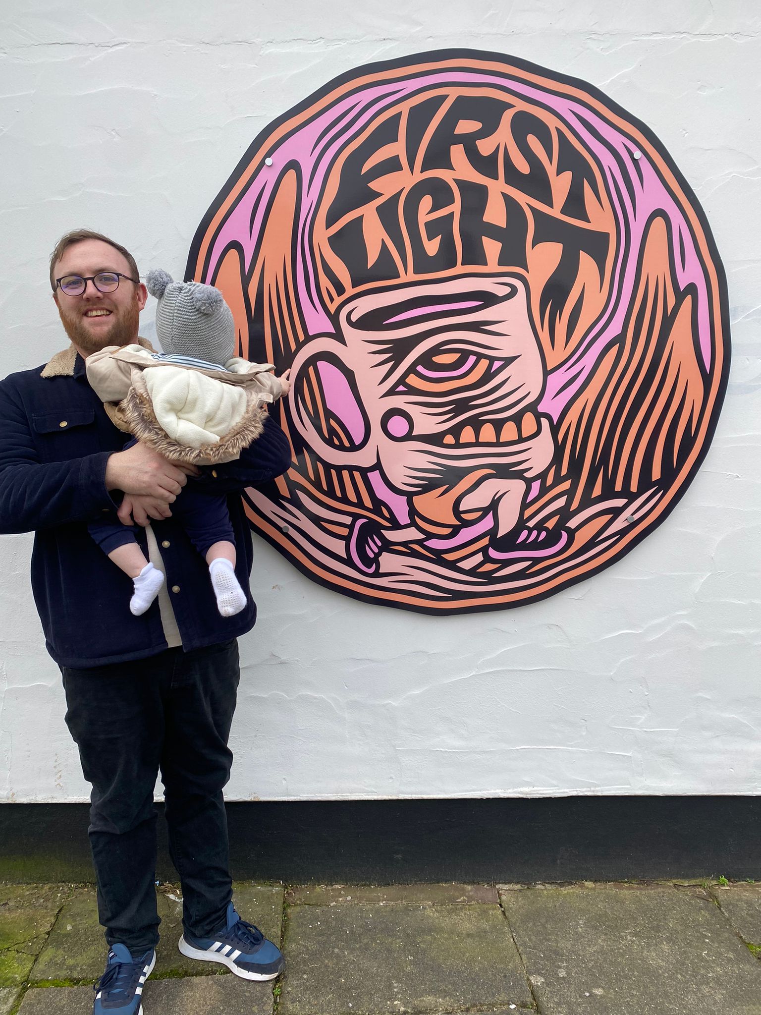 Jamie Green holds a baby in front of a colourful mural featuring a stylised coffee cup with legs and the words “FIRST LIGHT,” set against a pink and orange mountainous background.