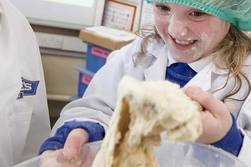 A smiling child wearing a hairnet and lab coat holds sticky dough in a bowl, with flour on her face in a classroom or kitchen setting.
