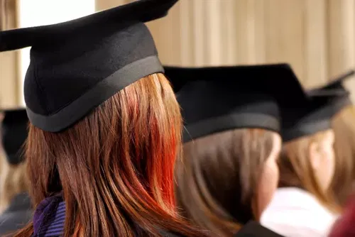 A close-up of students wearing black graduation caps and gowns, seated in rows, facing forwards during a graduation ceremony. The focus is on the back of a student with auburn hair.