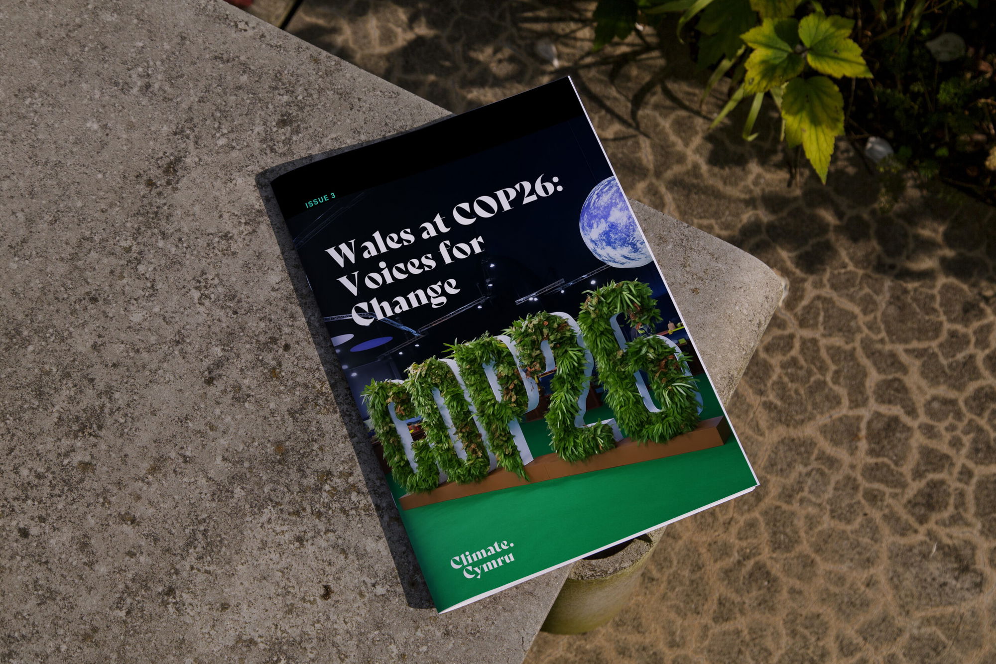 A magazine titled Wales at COP26: Voices for Change with greenery-covered COP26 letters and an Earth image on the cover rests on a concrete surface near green plants.