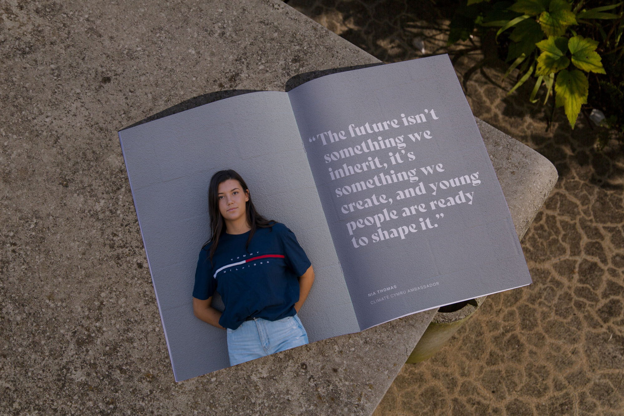 An open magazine on a stone surface shows a young woman in a blue shirt on one page and a quotation about the future and young people on the other, with plants and textured ground visible nearby.