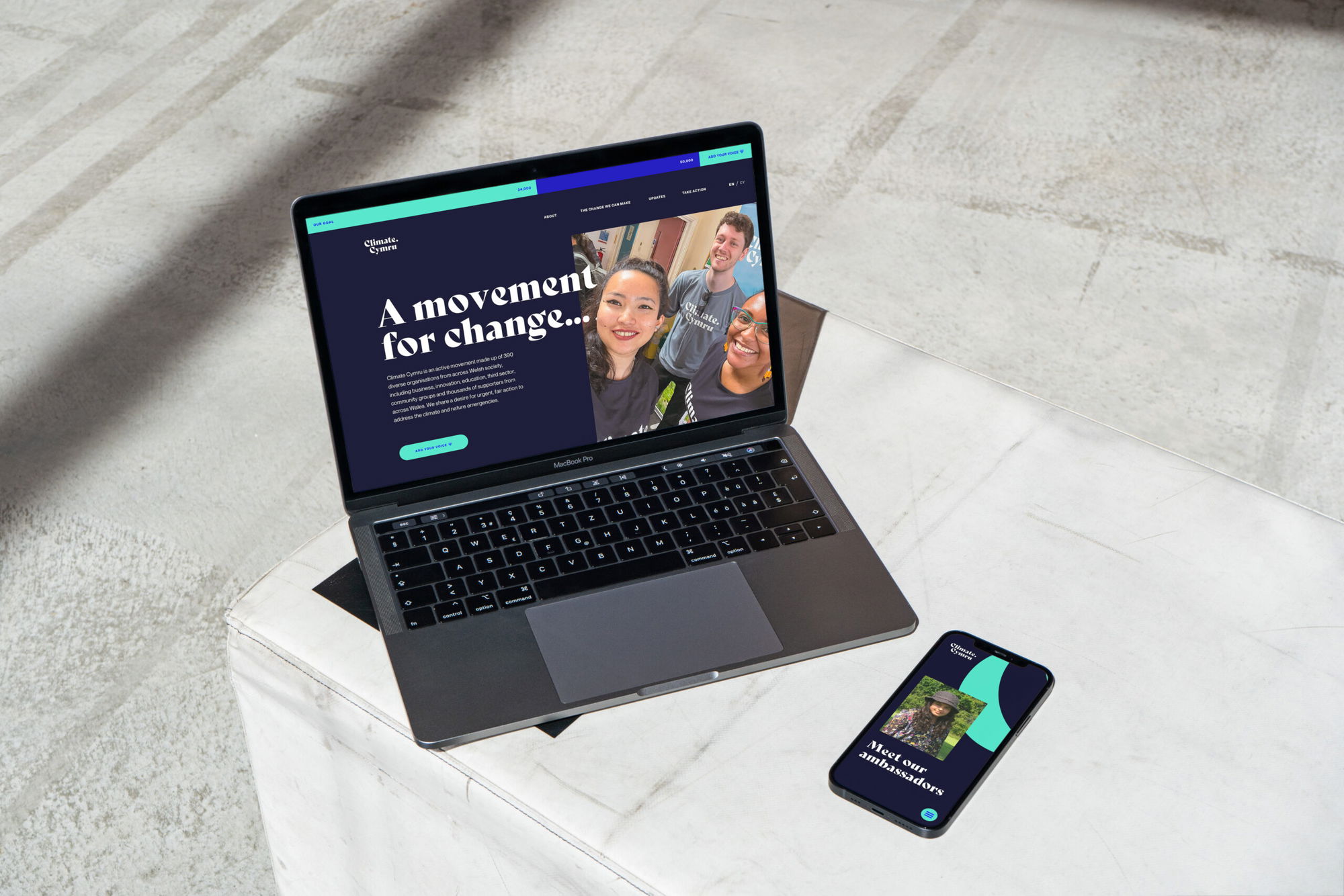A laptop and a smartphone display the same colourful website with the headline “A movement for change…” and images of smiling people, both resting on a white marble table in a bright, modern space.