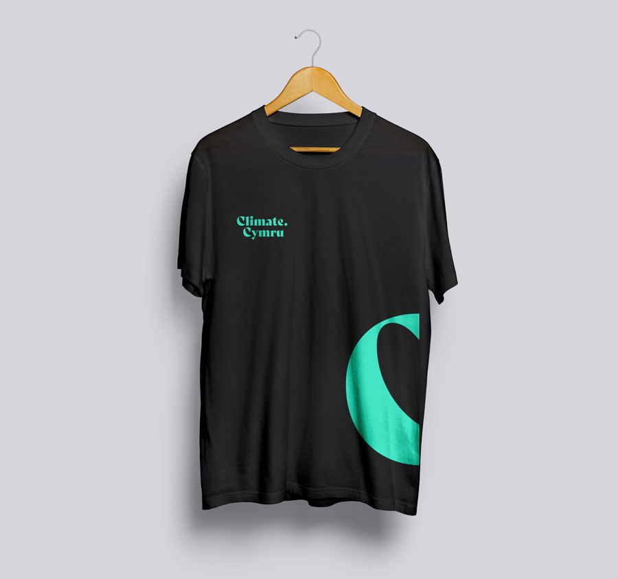 A black T-shirt on a wooden hanger features the text Climate. Cymru in turquoise on the left chest and a large turquoise letter C design on the lower right side. The background is plain light grey.