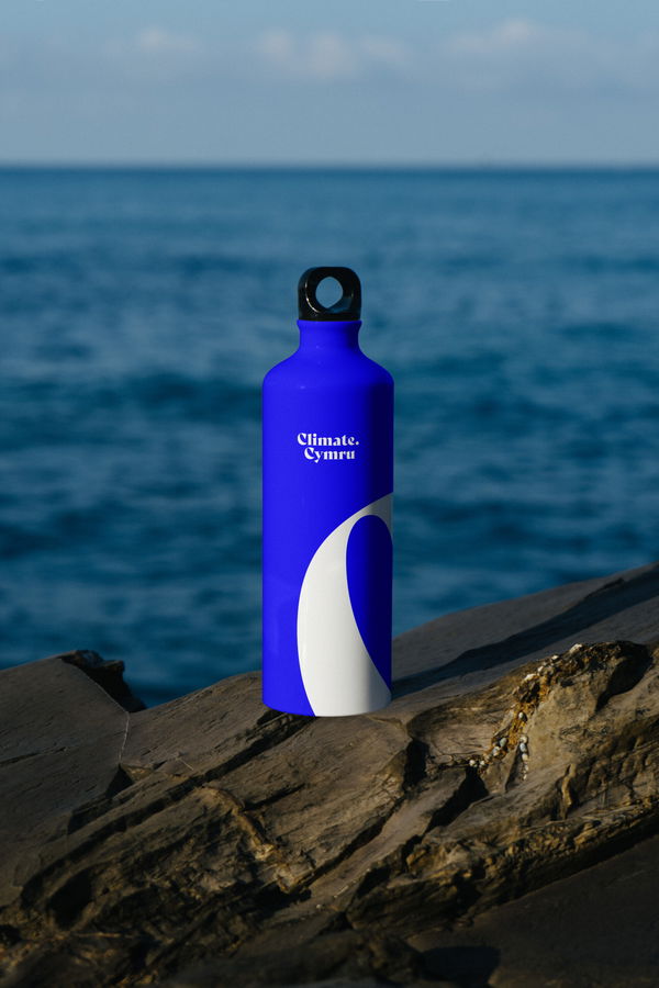 A blue reusable water bottle with a white design and the words Climate Cymru printed on it sits on a rocky surface, with the sea and sky blurred in the background.