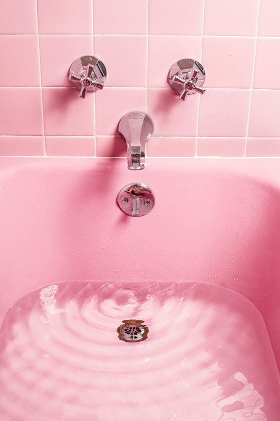 A pink bath with water running near the drain, chrome tap and knobs, pink tiles on the wall, and a dark blue tube of cream on the bath’s edge.