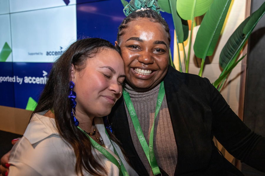 Two women with green lanyards smile warmly; one has her eyes closed and leans affectionately on the other’s shoulder, standing in front of a screen with “accent” partially visible and large green leaves in the background.