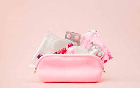 A pink pouch filled with sanitary towels, blister packs of pills, tampons, and wrapped sanitary products against a light pink background.