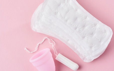 A sanitary towel, a white tampon, and a pink menstrual cup are arranged on a light pink background.