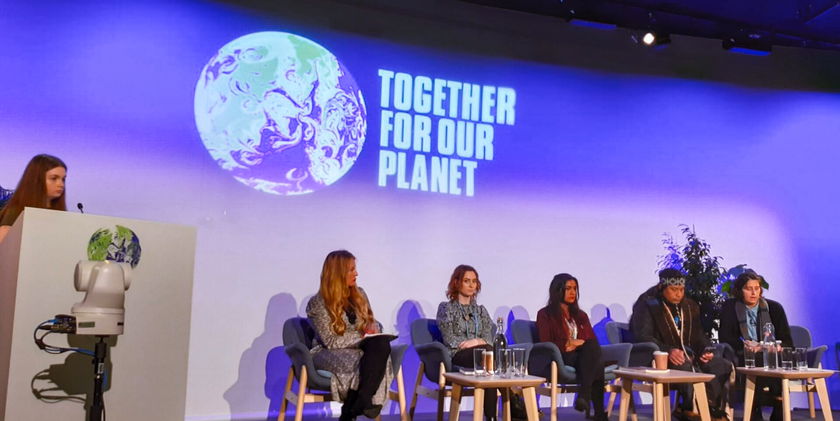 A panel of five people sits on stage beneath a TOGETHER FOR OUR PLANET sign and Earth graphic, while a woman speaks at a lectern on the left; glasses and water jugs are on the table in front.