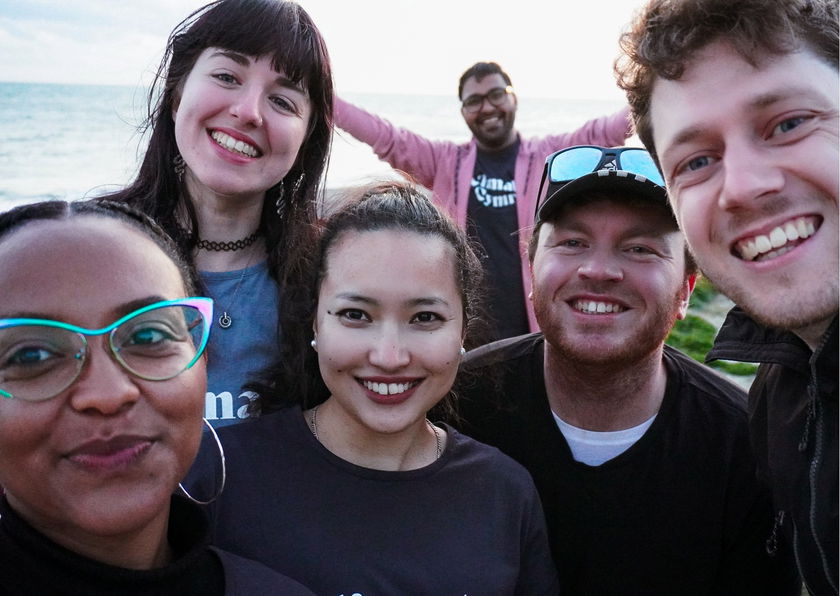 Six people smile and pose closely together for a group selfie outdoors by the water, with the sea and sky in the background. They appear happy and are dressed casually.